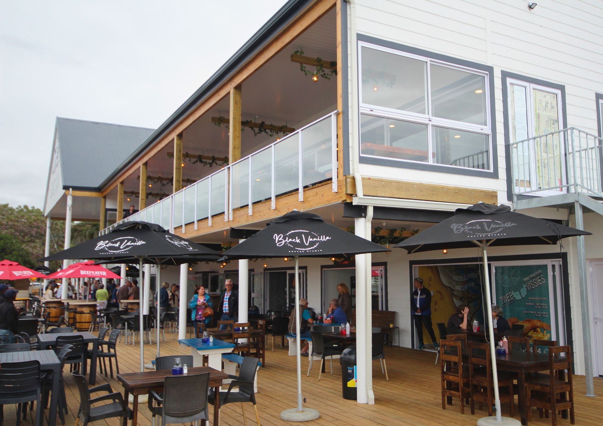 Outdoor seating area of a two-story restaurant with wooden deck, tables, chairs, and black umbrellas labeled Black Vanilla, with people dining and talking under an overcast sky.